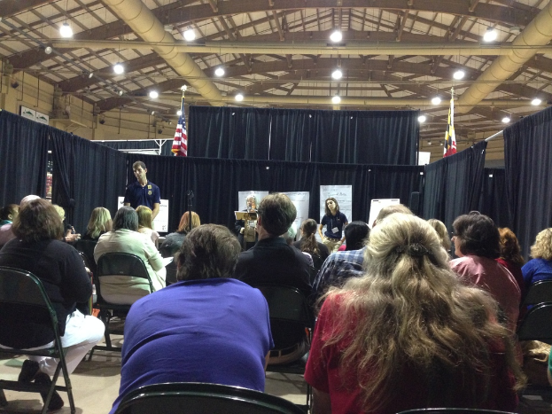 Eine Gruppe von Menschen, die auf Stühlen sitzen, mit einer Person, die an einem Rednerpult steht, vor einem Hintergrund aus Transparenten, blauen Vorhängen, Flaggen und Deckenleuchten.