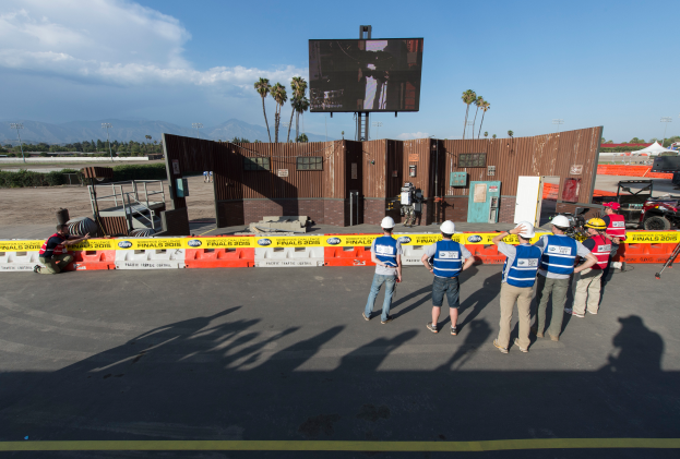 Eine Gruppe von Menschen in Helmen steht vor einem großen Bildschirm auf dem Las Vegas Motor Speedway, umgeben von Barrieren, Fahrzeugen, Hütten, Bäumen, Lichtmasten und Bergen mit einem bewölkten Himmel im Hintergrund.