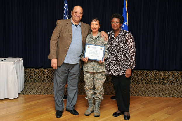 Mehrere Personen in formeller Kleidung stehend, wobei eine Person in der Mitte ein Zertifikat hält, Flaggen vor Vorhängen und ein linksseitiger, mit einem Tuch bedeckter Tisch.