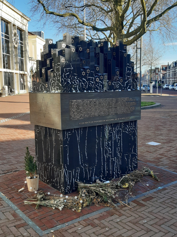 Denkmal in einem Stadtplatz, das Holocaust-Opfern gewidmet ist, mit einem Baum davor, einem Topfgewächshaus auf dem Boden, verstreuten trockenen Blättern und im Hintergrund Gebäude, Fahrzeuge, Mäste und Bäume.