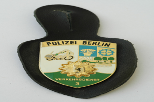 Ein schildförmiges Polizeibadged mit "Polizei Berlin" in schwarzer fettgedruckter Schrift auf einem blauen Hintergrund mit einem weißen Stern und Rahmen, das auf einer Oberfläche liegt.