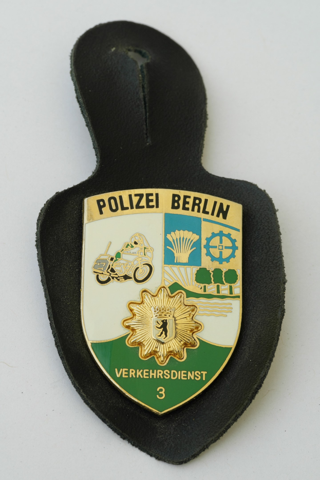Ein schildförmiges Polizeibadged mit "Polizei Berlin" in schwarzer fettgedruckter Schrift auf einem blauen Hintergrund mit einem weißen Stern und Rahmen, das auf einer Oberfläche liegt.