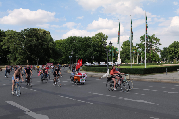 Eine Gruppe von Menschen, die auf Fahrrädern eine von Bäumen gesäumte Straße mit Laternenmasten und Fahnenmasten entlangfahren, während der Paris Bike Ride 2016, mit Fahrzeugen im Hintergrund und einem bewölkten Himmel.