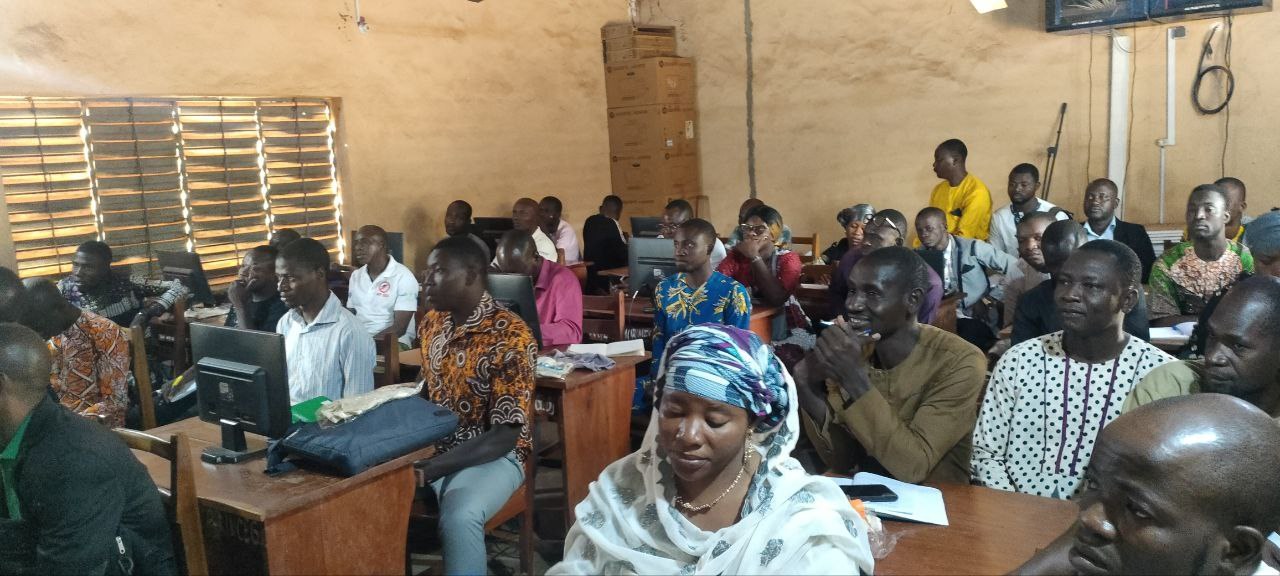 Eine Gruppe von Menschen sitzt an Tischen in einem Klassenzimmer mit Monitoren, Taschen, Büchern und Papieren und nimmt an einem Techniktraining teil.