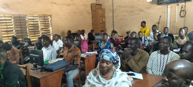 Eine Gruppe von Menschen sitzt an Tischen in einem Klassenzimmer mit Monitoren, Taschen, Büchern und Papieren und nimmt an einem Techniktraining teil.