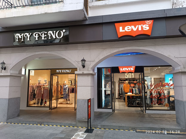 Storefront of Levi's clothing brand with mannequins in dresses, hanging clothes, illuminated displays, and text boards, situated above a road.
