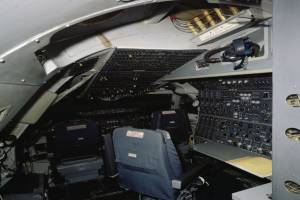 Ein Flugzeugcockpit mit zwei Sitzen, einem Tisch mit einem Buch darauf und verschiedenen elektronischen Geräten und Kabeln.