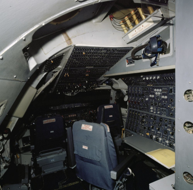 Ein Flugzeugcockpit mit zwei Sitzen, einem Tisch mit einem Buch darauf und verschiedenen elektronischen Geräten und Kabeln.