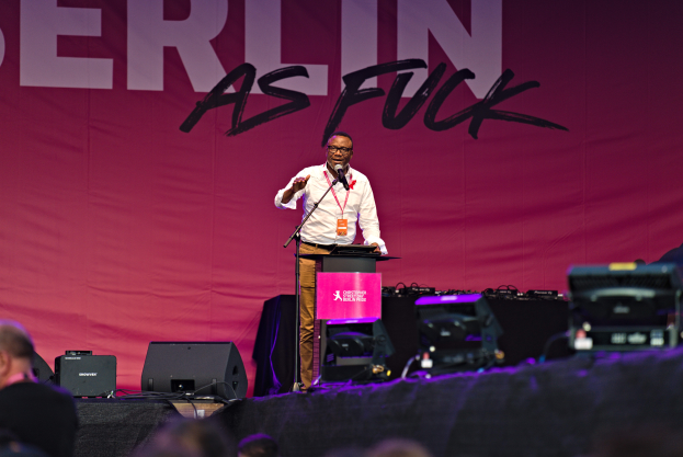 Mann in weißem Hemd und braunen Hosen steht auf einer Bühne mit Mikrofon und pinkem Banner 'Berlin as Fuck'