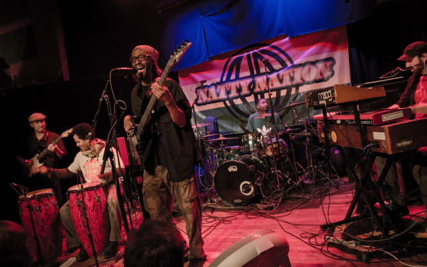 Eine Live-Band performt auf der Bühne mit Mikrofonen und Ständern, während das Publikum unten zuschaut; im Hintergrund ist ein Banner mit Text zu sehen.