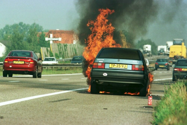 Ein Auto in Flammen auf dem Seitenstreifen mit anderen Fahrzeugen in der Nähe, umgeben von Bäumen, Gebäuden und einem klaren blauen Himmel, mit Gras und einem Feuerlöscher auf der rechten Seite.