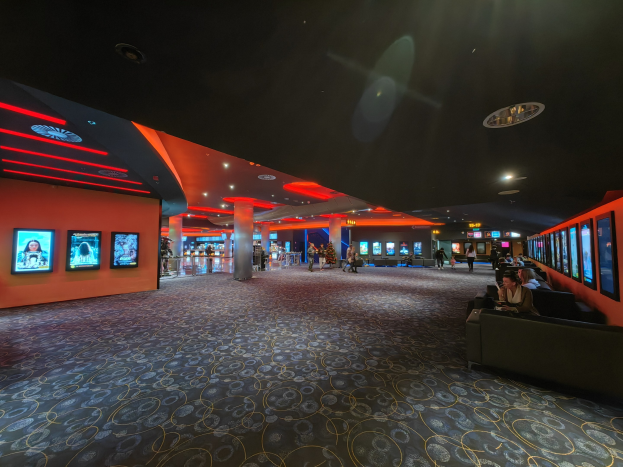 Ein geräumiger Raum mit Menschen auf Sofas sitzend, die an der Wand montierten Fernseher beobachten, umgeben von Säulen und Deckenleuchten, mit einem sichtbaren Boden.