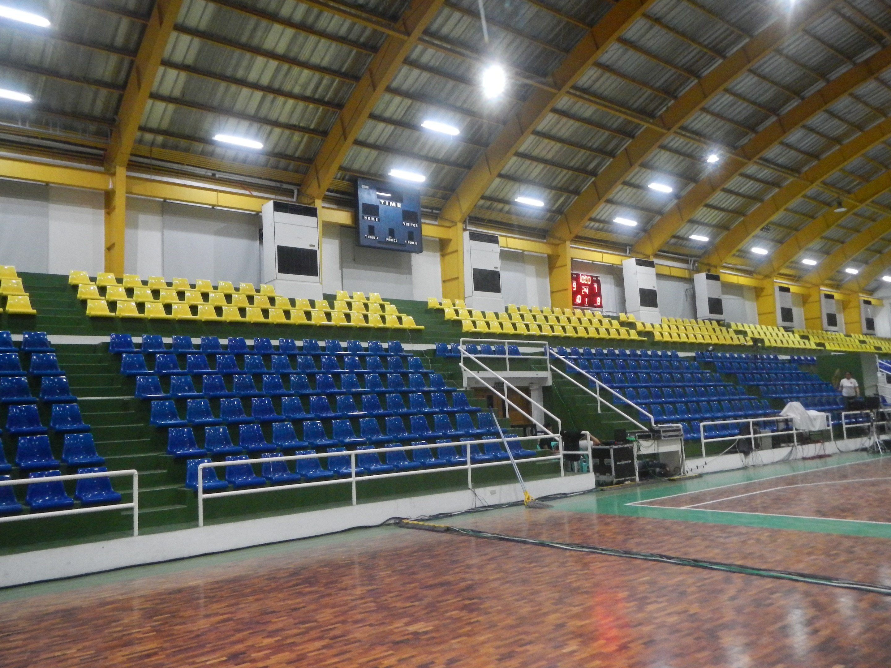 Großer Hallensportplatz mit Holzboden, blauen und gelben Sitzplätzen, Geländern, Treppen zu den Sitzplätzen, Wandbildschirme, Deckenleuchten und einigen Menschen.