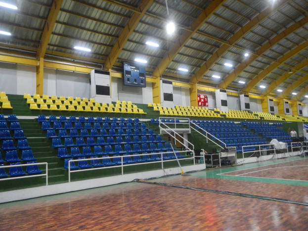 Großer Hallensportplatz mit Holzboden, blauen und gelben Sitzplätzen, Geländern, Treppen zu den Sitzplätzen, Wandbildschirme, Deckenleuchten und einigen Menschen.