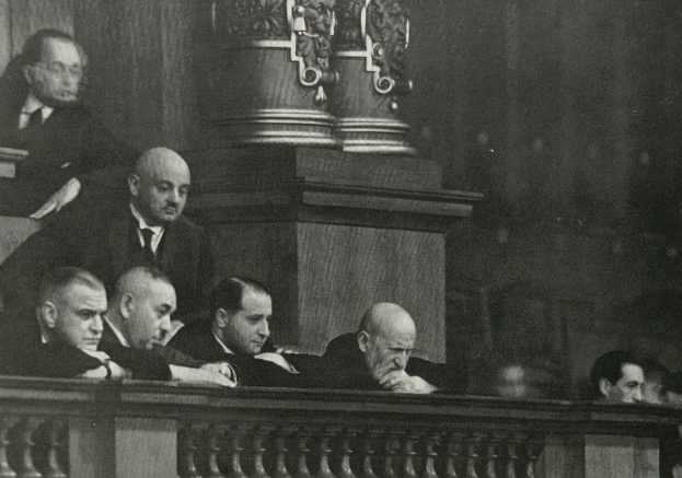 Eine Gruppe von M├Ąnnern sitzt in einem schwarz-wei├čen Gerichtssaal, mit einem h├Âlzernen Gel├Ąnder im Vordergrund und einer S├Ąule im Hintergrund, w├Ąhrend eines Prozesses.