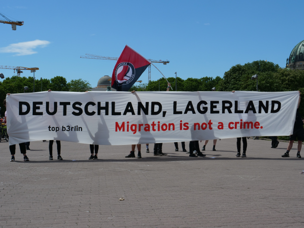 Eine Gruppe von Menschen mit einem Banner, auf dem "Deutschland, Lagerland Migration ist kein Verbrechen" steht, und einer Flagge, mit Straßenlaternen, Bäumen, Kränen, einem Gebäude und einem bewölkten Himmel im Hintergrund.