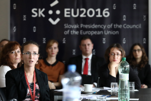 Eine Gruppe von Menschen sitzt um einen Tisch mit Wasserflaschen, Gläsern, Tassen, Untertäsern, Papieren, Stiften und anderen Gegenständen während einer scheinbaren Sitzung der Europäischen Union, mit einem Banner und einer Wand im Hintergrund.