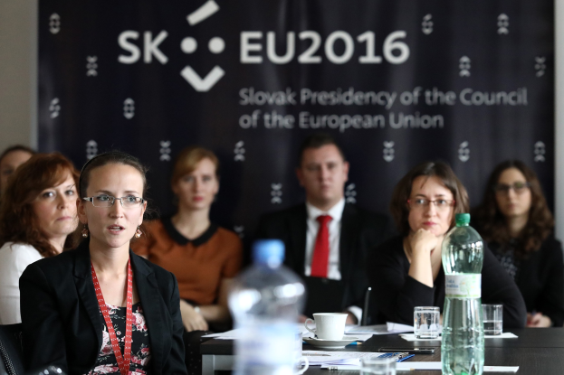 Eine Gruppe von Menschen sitzt um einen Tisch mit Wasserflaschen, Gläsern, Tassen, Untertäsern, Papieren, Stiften und anderen Gegenständen während einer scheinbaren Sitzung der Europäischen Union, mit einem Banner und einer Wand im Hintergrund.