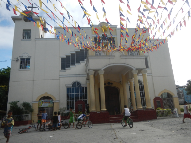 Eine Gruppe von Menschen auf Fahrrädern vor einer weißen Kirche mit architektonischen Details, umgeben von Grün, einem Banner, Bäumen und einem bewölkten Himmel, mit bunten Fahnen über einem Seil.