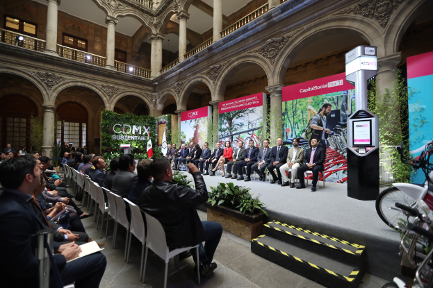 Eine Gruppe von Menschen, die auf Stühlen sitzen und einem Mann auf einer Bühne gegenübersitzen, der ein Mikrofon hält, mit Fahrrädern rechts geparkt und verschiedenen Elementen wie Plakaten, Pflanzen, Säulen, Geländern, Lampen, Fenstern und einer Wand im Hintergrund.