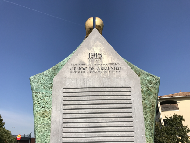 Ein steinernes Denkmal mit einer Uhr darauf, umgeben von Bäumen und Gebäuden, mit einer Inschrift, die den 24. Jahrestag des Völkermords in Armenien erinnert.