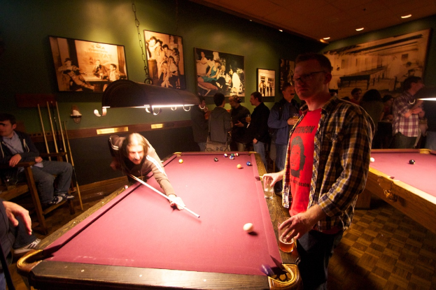 In einem Club spielen zwei Personen Snooker an einem von zwei Tischen, während mehrere andere im Hintergrund stehen und sich unterhalten, vor einer cremefarbenen Wand mit ein paar Fotorahmen.