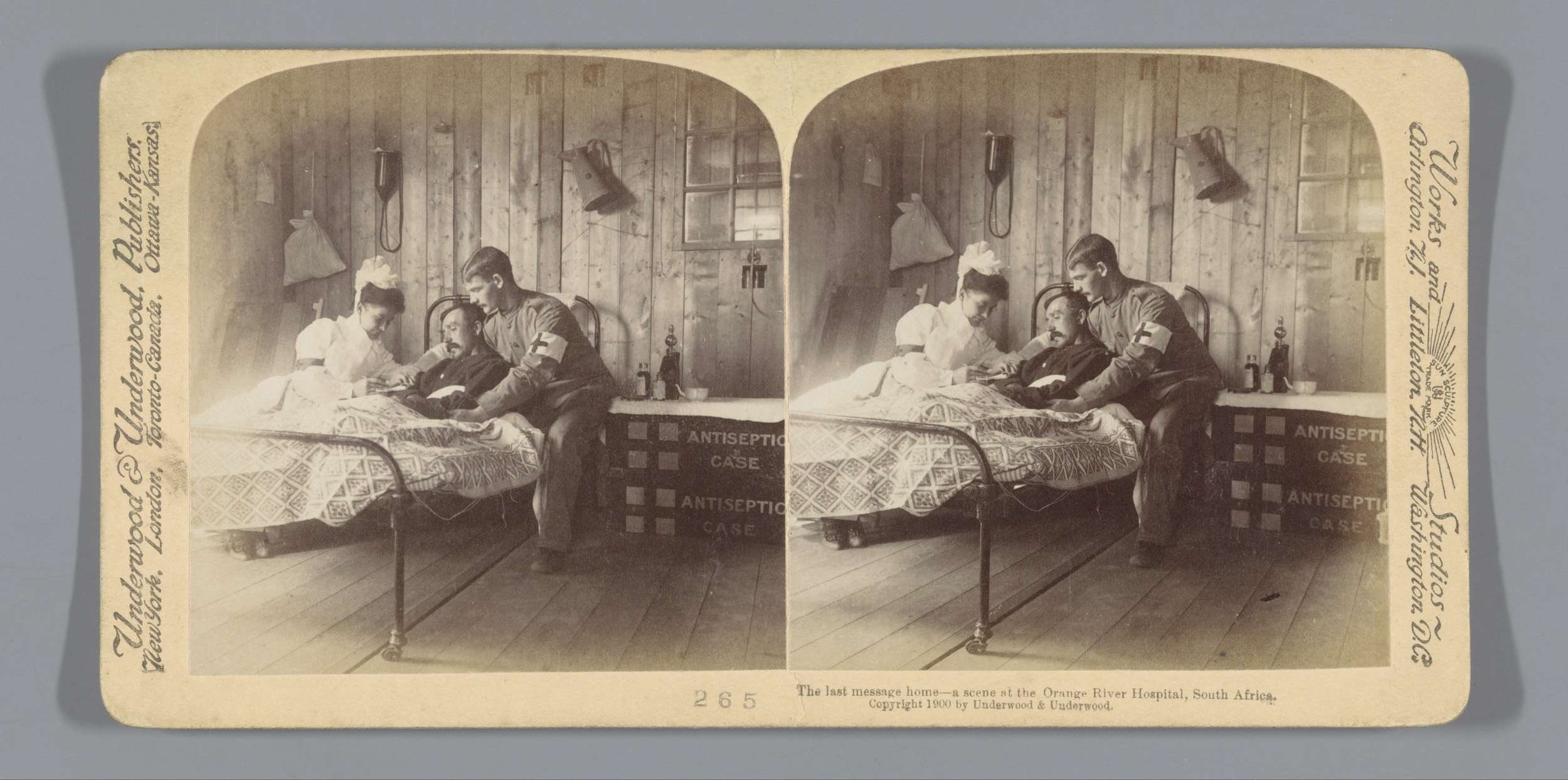 Ein altes Schwarz-Weiß-Foto eines Krankenhauszimmers mit zwei Patienten im Bett, zwei stehenden Personen und einem Tisch mit Gegenständen, mit Wandlampen und einem Fenster, mit Text unten mit der Aufschrift "Die letzte Botschaft nach Hause - eine Szene am Orange River Hospital, Südafrika".