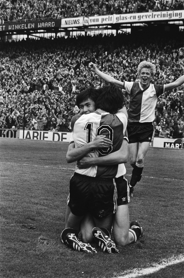 Drei Fußballspieler umarmen sich auf dem Boden nach einem Torerfolg, mit jubelnder Menge und schwenkenden Fahnen in einem schwarz-weißen Stadion Setting.