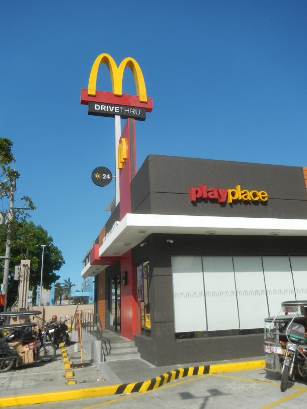 Ein McDonald's Restaurant mit einem "Playplace"-Schild an der Seite, umgeben von Fahrzeugen, B├Ąumen, Masten und einem klaren blauen Himmel.