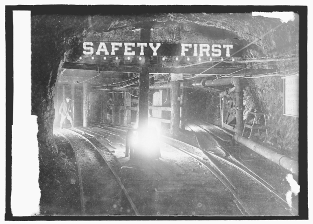 Schwarz-weißes Foto eines aus einem Tunnel fahrenden Zuges mit einer Person auf den Gleisen im Vordergrund, Hintergrund zeigt Strommasten, Lampen und ein "Sicherheit zuerst"-Schild.