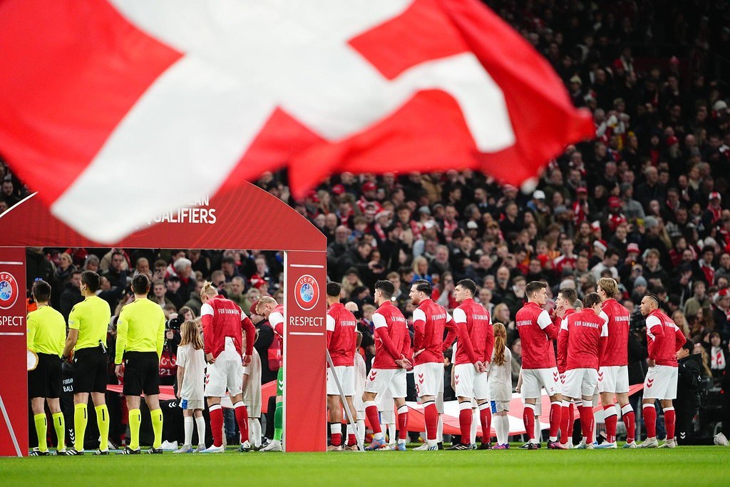Gruppe von Menschen auf einem Fußballfeld mit einer roten und weißen Flagge im Vordergrund, einem Bogen mit der Aufschrift "Bayern München vs Bayern Munich Betting Tips & Preview" im Hintergrund und einer großen Menge im Stadion.