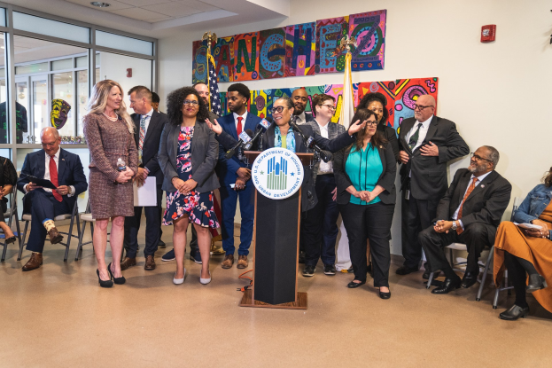 Gruppe von Menschen vor einem Rednerpult mit einem Mikrofon und einer Tafel mit der Aufschrift "Sancho Stadtrat joins City Council", mit sitzenden und stehenden Teilnehmern, Flaggen, Feuerlöscher und Deckenleuchten.