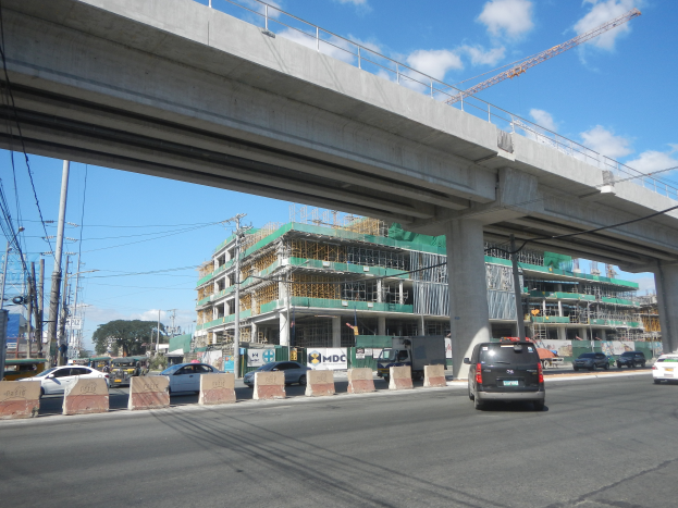 Eine belebte Hauptstraße mit Fahrzeugen, einer Brücke, Strommasten, Baustellen, Bäumen und einem bewölkten Himmel.