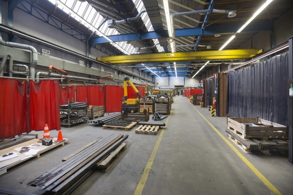 Großes Lagerinnere mit gestapelten Holzpaletten, verstreuten Metallobjekten, Verkehrskegeln und einem zentralen Maschinenfahrzeug, mit roten Vorhängen auf der linken Seite, einer Vorhangwand auf der rechten Seite und Überkopfrohren und -leuchten.