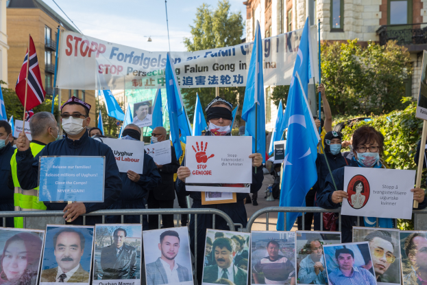 Eine Gruppe von Menschen mit Masken und Fahnen und Schildern steht vor einem Zaun mit Fotos und Text, protestiert in Istanbul, Türkei, mit Gebäuden, Bäumen und einem Laternenpfahl im Hintergrund.
