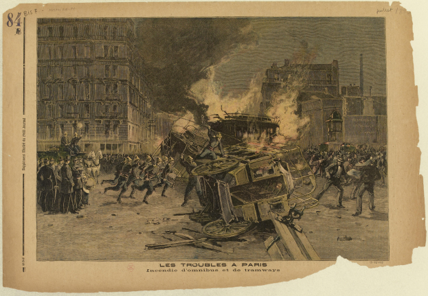 Eine Gruppe von Menschen steht um ein brennendes Auto in der Mitte einer Straße, mit Gebäuden im Hintergrund und Text unten im Bild.