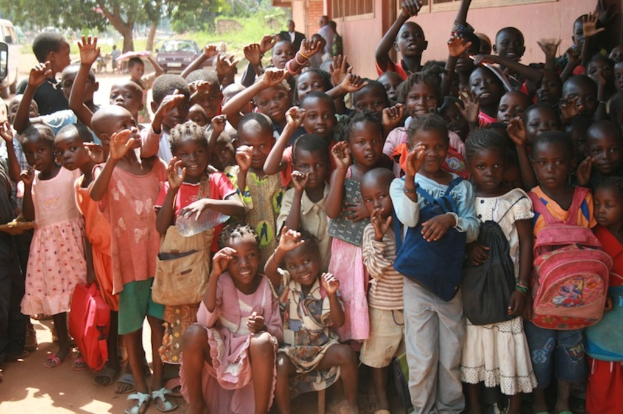 Eine Gruppe von Kindern, die mit erhobenen Händen stehen, mit einem Gebäude auf der rechten Seite und einem Fahrzeug unter Bäumen im Hintergrund.