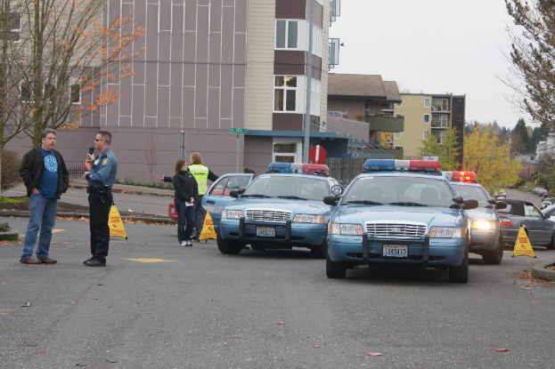 Autos auf einer Straße mit vier Menschen in der Nähe, Gebäude mit Fenstern im Hintergrund, Bäume und Warnbörsen.