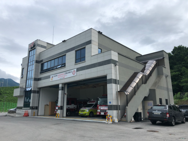 Eine Feuerwache mit parkenden Autos davor, mit einer Treppe, die zum Eingang führt, einem Schild und verstreuten Verkehrskegeln, mit Bäumen, Hügeln und einem bewölkten Himmel im Hintergrund.