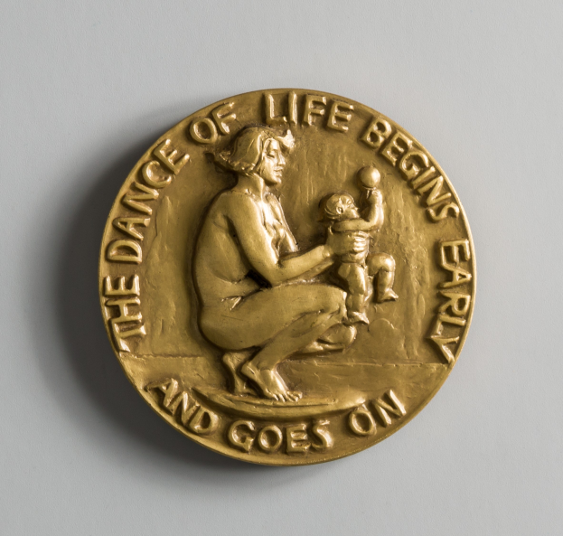 Eine Goldmedaille mit der Inschrift "Der Tanz des Lebens beginnt früh und geht weiter" und ein Bild einer Frau mit einem Baby.
