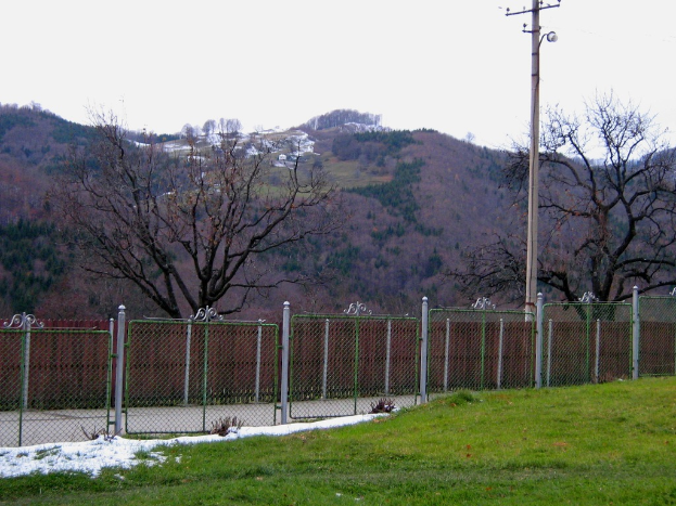 Eine grüne Landschaft mit einem Zaun und einem Pfahl im Vordergrund, Bäume, die über das Gebiet verstreut sind, und einige Häuser, die auf einem Hügel in der Ferne zu sehen sind.