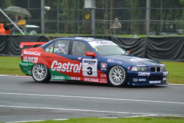 Ein Auto fährt auf einer Rennstrecke mit Zuschauern hinter einem Maschendrahtzaun, einige halten Schirme, mit Gras auf beiden Seiten und Bäumen in der Ferne.