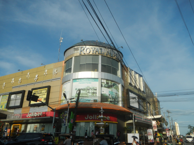 Eine belebte Stadtstraße mit fahrenden Fahrzeugen, Fußgängern auf dem Gehweg und dem Royal Mall Einkaufszentrum im Hintergrund, umgeben von parkenden Autos, Bäumen, Strommasten und Drähten unter dem Himmel.