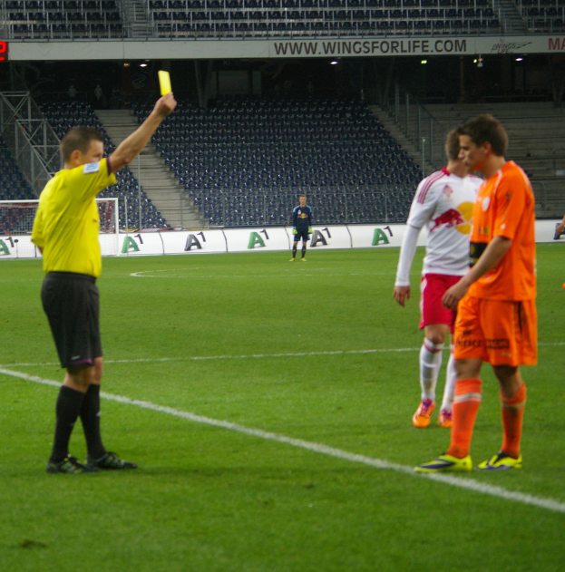 Ein Schiedsrichter zeigt einem Fussballspieler auf dem Feld eine gelbe Karte, umgeben von einer Gruppe von Menschen, mit einem Torpfosten, einem Zaun, einer Tafel, Metallrahmen, einem Anzeigebildschirm, Stühlen und einer Decke mit Deckenleuchten im Hintergrund.