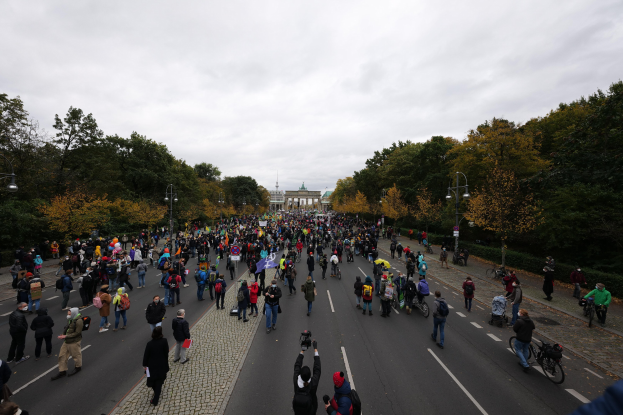 Eine große Gruppe von Menschen marschiert auf einer Straße mit Bäumen und Laternen in Berlin, einige halten Kameras und ein Gebäude ist im Hintergrund unter einem klaren Himmel zu sehen.