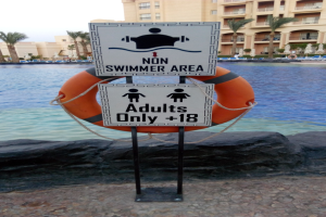 Ein Schwimmbad mit einem "Nur für Erwachsene - Nicht Schwimmer Bereich"-Schild im Vordergrund, umgeben von Bäumen und Gebäuden unter einem klaren blauen Himmel, mit einem Rettungsring in der Mitte des Pools.