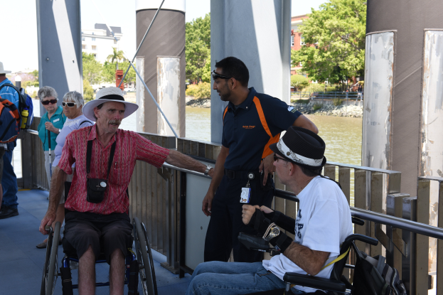 Eine Gruppe von Menschen auf einer Brücke mit zwei Rollstuhlfahrern im Vordergrund, umgeben von einem Geländer, mit einem Gewässer, Bäumen, Gebäuden und einem klaren blauen Himmel im Hintergrund.