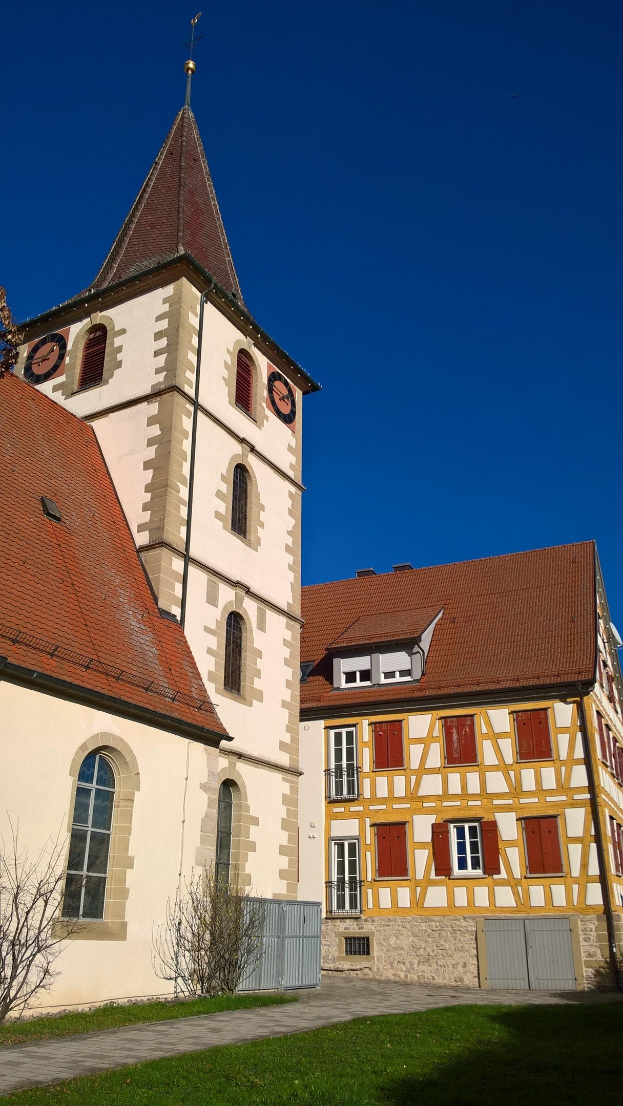 Eine Kirche mit einem zentralen Turm und einem Uhrenturm, umgeben von Gebäuden, einem Metallzaun, einem Weg, Pflanzen, Gras und einem bewölkten Himmel.