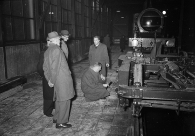 Eine Gruppe von Männern in Hüten steht um einen Zug in einer Fabrik herum, wobei einer der Männer auf dem Boden sitzt, der Zug rechts und eine Wand links zu sehen ist, dargestellt in Schwarz-Weiß.