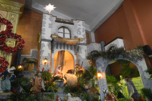 Eine Krippe in einer Kirche, die für Weihnachten geschmückt ist, mit Statuen von Menschen, Tieren, Pflanzen, Lichtern und anderen Gegenständen und einem Stern oben und einer Wand im Hintergrund.